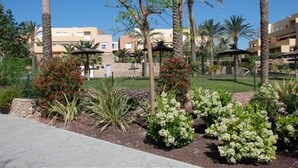 A landscaped garden with palm trees, a pathway, and buildings in the background. - Holiday Home "Los Almendros" with Shared Pool, Terrace & Wi-Fi (Vera)
