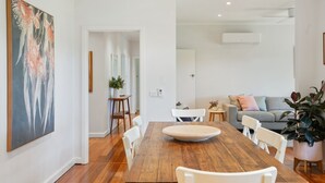 Uma sala de jantar com mesa de madeira e cadeiras brancas, um sofá cinzento e um quadro abstrato grande na parede.