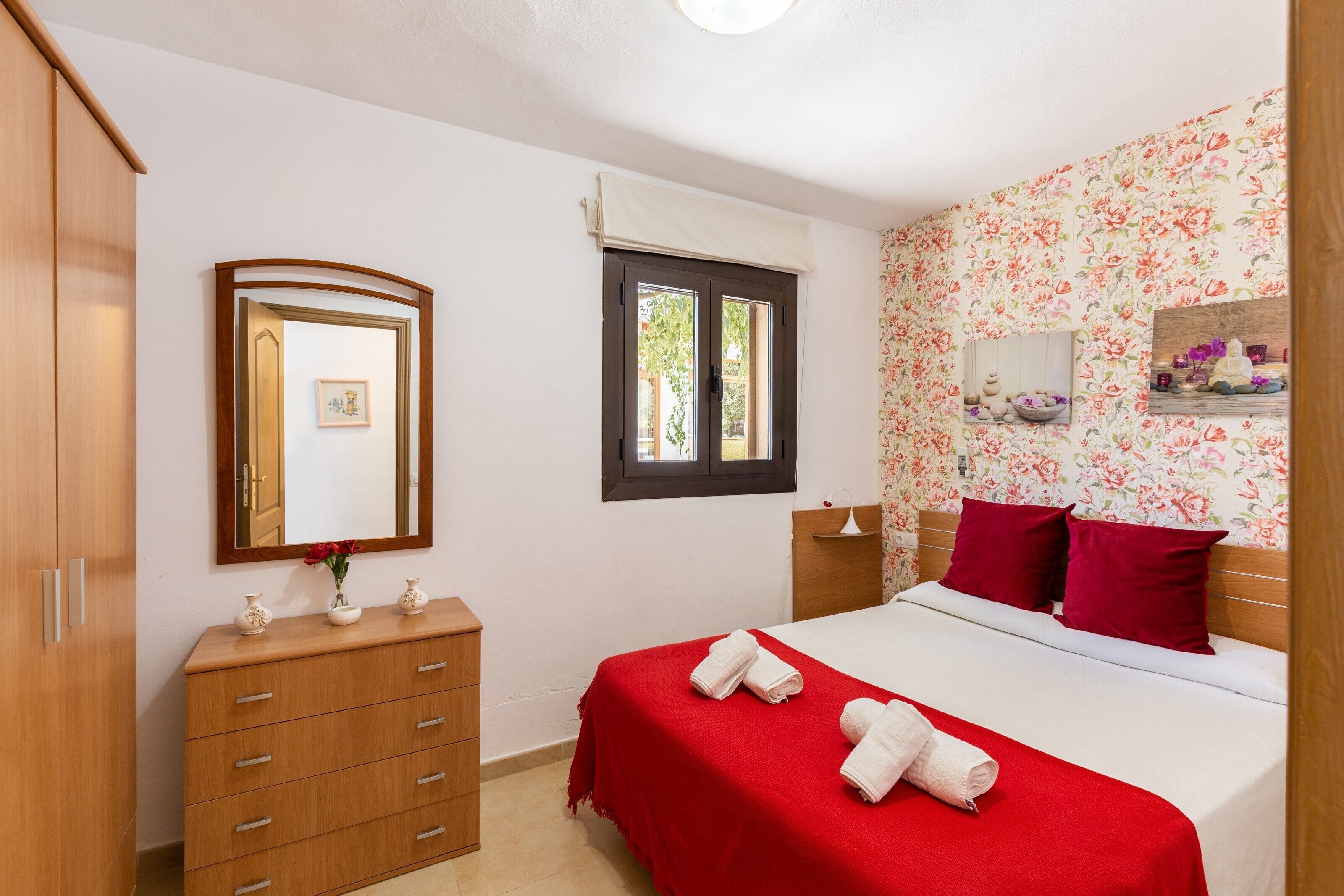 Une chambre à coucher avec un lit, une commode, un miroir, une fenêtre et un papier peint à motifs floraux.