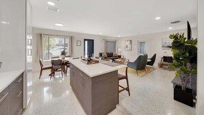 Una cocina moderna con una isla central, comedor con sillas y sala de estar con sofá y mesa de centro.
