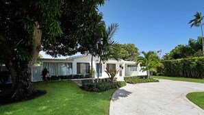 Una casa moderna de dos plantas con un camino de entrada curvo, rodeada de exuberante follaje y palmeras.