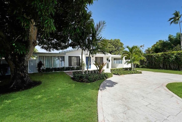 A modern two-story house with a curved driveway, surrounded by lush greenery and palm trees.