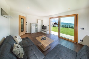 A modern living room with a sofa, a coffee table, and a sliding glass door leading to a balcony with a view of the mountains.