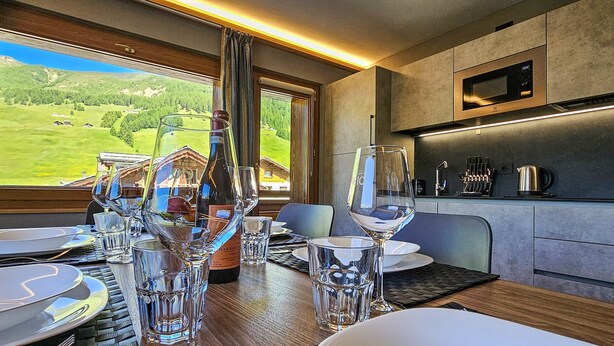 Een eethoek met een tafel voor twee, een fles wijn en uitzicht op een bergachtig landschap door een raam.