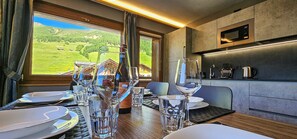Un comedor con una mesa dispuesta para dos personas, una botella de vino y vistas a un paisaje montañoso a través de una ventana.