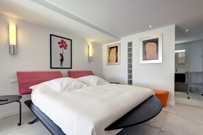 A modern hotel room with a bed, a small table, a red chair, and artwork on the walls.