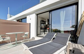 A modern outdoor lounge area with two reclining chairs, a small table, and a glass door leading to a kitchen.