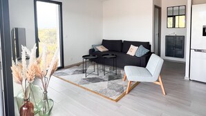 Una sala de estar moderna con un sofá, una mesa de centro y una silla. Hay un ventanal y una puerta que da a otra habitación.