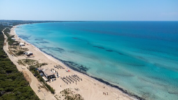 An aerial view of a beach with clear turquoise waters, a sandy shore, and a few buildings along the coastline.