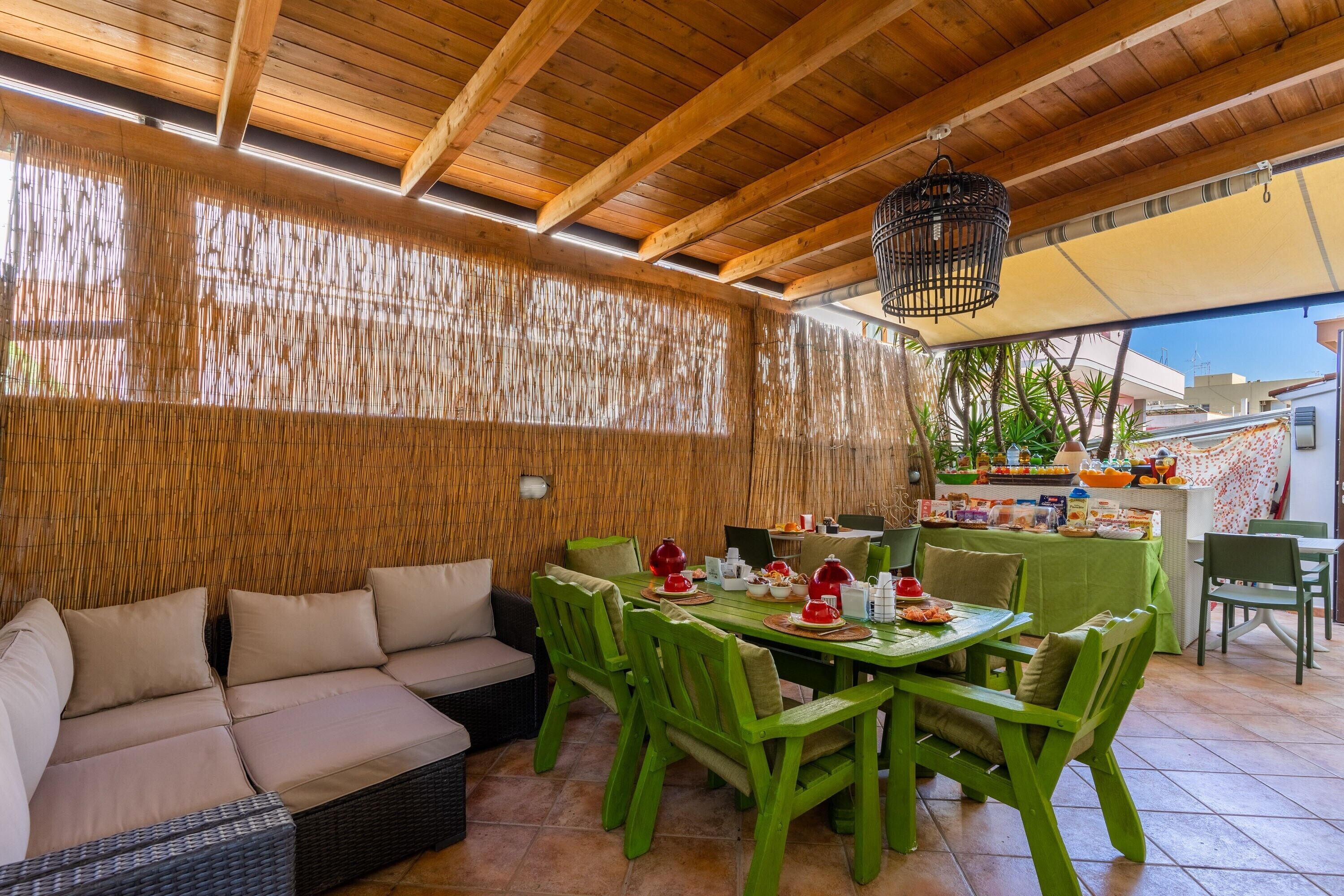 An outdoor dining area with a wooden ceiling, green chairs, a table set with dishes, and a wicker sofa.