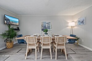 Un coin repas avec une table en bois, quatre chaises en osier, un téléviseur fixé au mur et un tableau encadré.