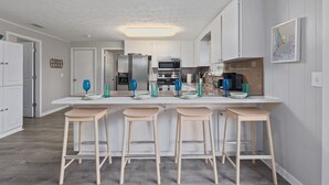 Una cocina moderna con armarios blancos, un refrigerador de acero inoxidable, un microondas y una encimera de granito. Hay cuatro taburetes de madera y cristalería azul sobre la encimera.