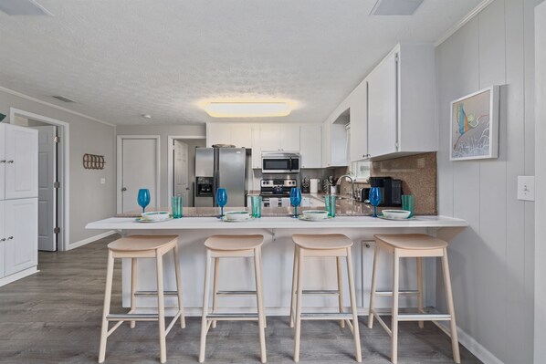 A modern kitchen with white cabinets, a stainless steel refrigerator, a microwave, and a granite countertop. There are four wooden stools and blue glassware on the counter. - Private Heated Pool Minutes to Mayo & Jax Beach (Jacksonville Beach)