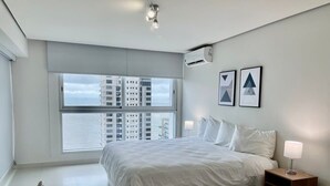 A modern bedroom with a large bed, a bedside table with a lamp, and a view of high-rise buildings through a large window.