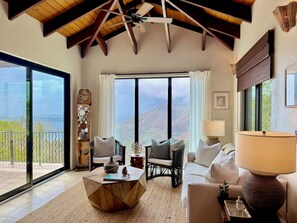 A spacious living area with a large window offering a view of the mountains, a ceiling fan, and a wooden coffee table. - Sapphire Peak 2br/2ba Private Pool, Panoramic Views, AC throughout, generator!!! (Coral Bay)
