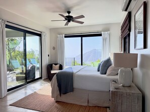Une chambre avec un grand lit, un ventilateur de plafond, une fenêtre avec des rideaux, une table de chevet avec une lampe et une vue sur l’extérieur.