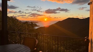 Vista al atardecer desde un balcón con mesa y sillas, con vistas a un cuerpo de agua y colinas.