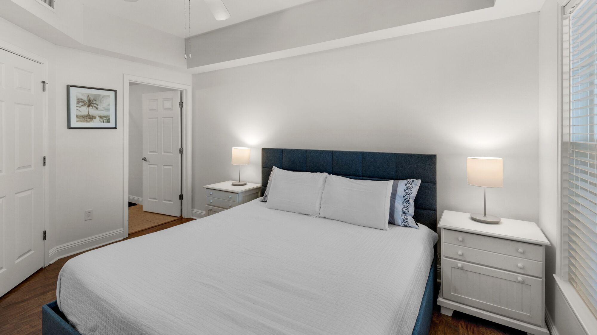 A bedroom with a large bed, two bedside tables with lamps, a door, and a framed picture on the wall.