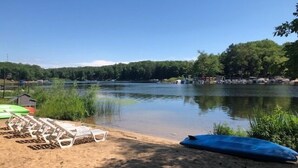 Blick auf einen See mit Booten, einen Sandstrand und bewaldetes Ufer im Hintergrund.