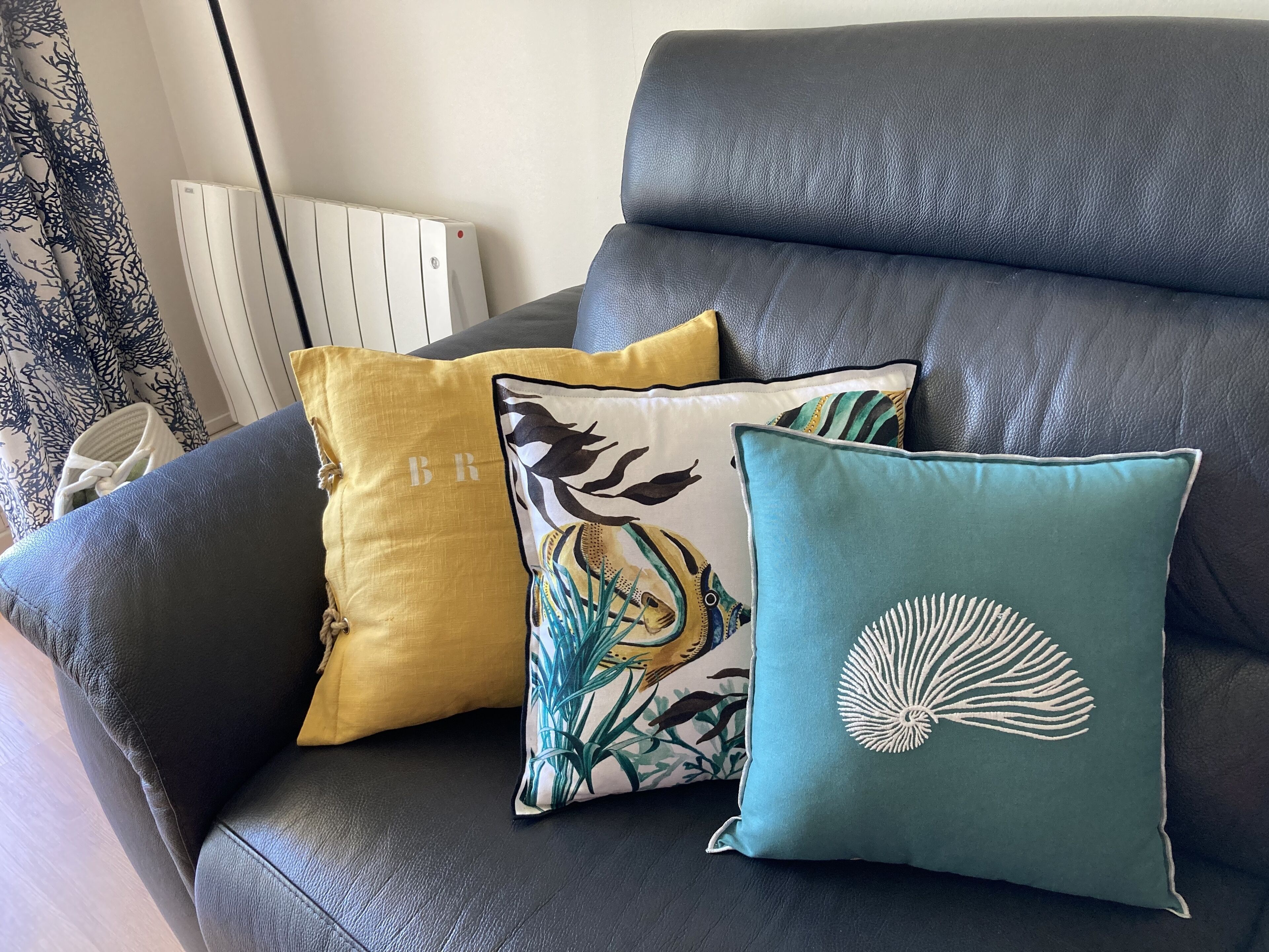 A black leather sofa with three decorative pillows, one with a bird design, one with a shell design, and one with a letter 'B' on it.