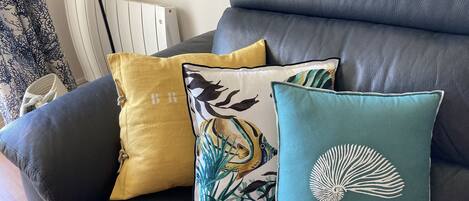 A black leather sofa with three decorative pillows, one with a bird design, one with a shell design, and one with a letter 'B' on it.
