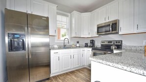 A modern kitchen with white cabinetry, stainless steel appliances, granite countertops, and a window with blinds. - 5105 The Retreat - Barefoot Resort Rentals (North Myrtle Beach)