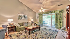 A cozy living room with a green sofa, a wooden coffee table, and a patterned rug. There is a ceiling fan, a window with curtains, and a view of the outdoors. - 533 Wedgewood - Barefoot Resort Rentals (North Myrtle Beach)