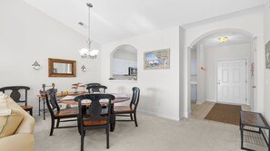 A dining area with a round table, chairs, and a chandelier. There is a kitchen visible in the background with white cabinets and a microwave.