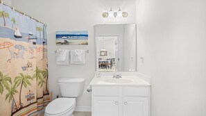 A bathroom with a beach-themed shower curtain, a toilet, a sink with a mirror, and a view into the adjacent dining area.