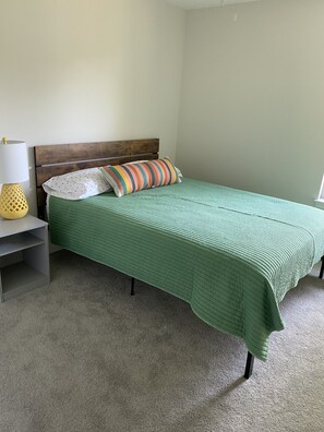 Un dormitorio con colcha verde acolchada, cabecera de madera, mesita de noche con lámpara amarilla y almohada blanca con estampado de lunares.