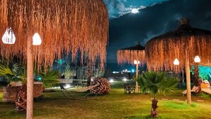A nighttime outdoor area with thatched umbrellas and string lights, surrounded by grass and palm trees. - ALBIZIA BOUTIQUE HOTEL (Ula)