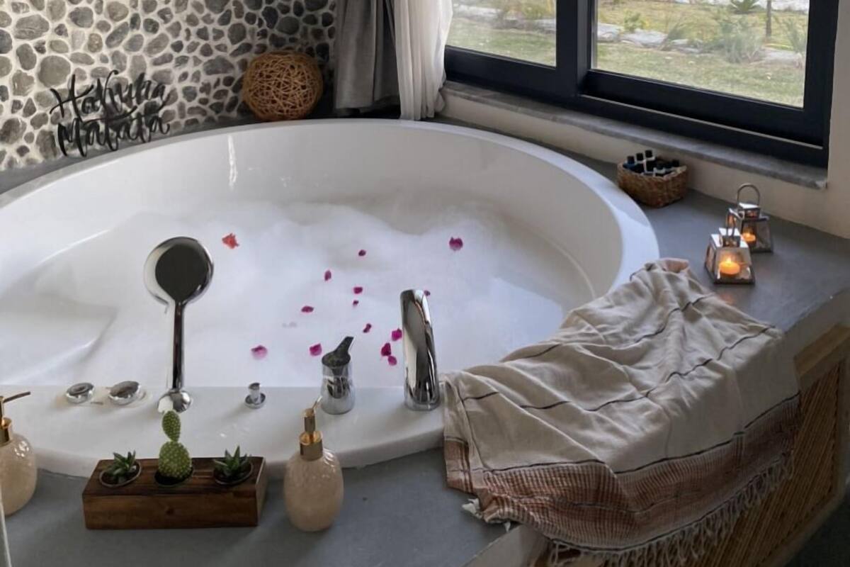 A bathroom with a stone wall, a large bathtub with rose petals, a window with a view of a landscape, and a towel rack.