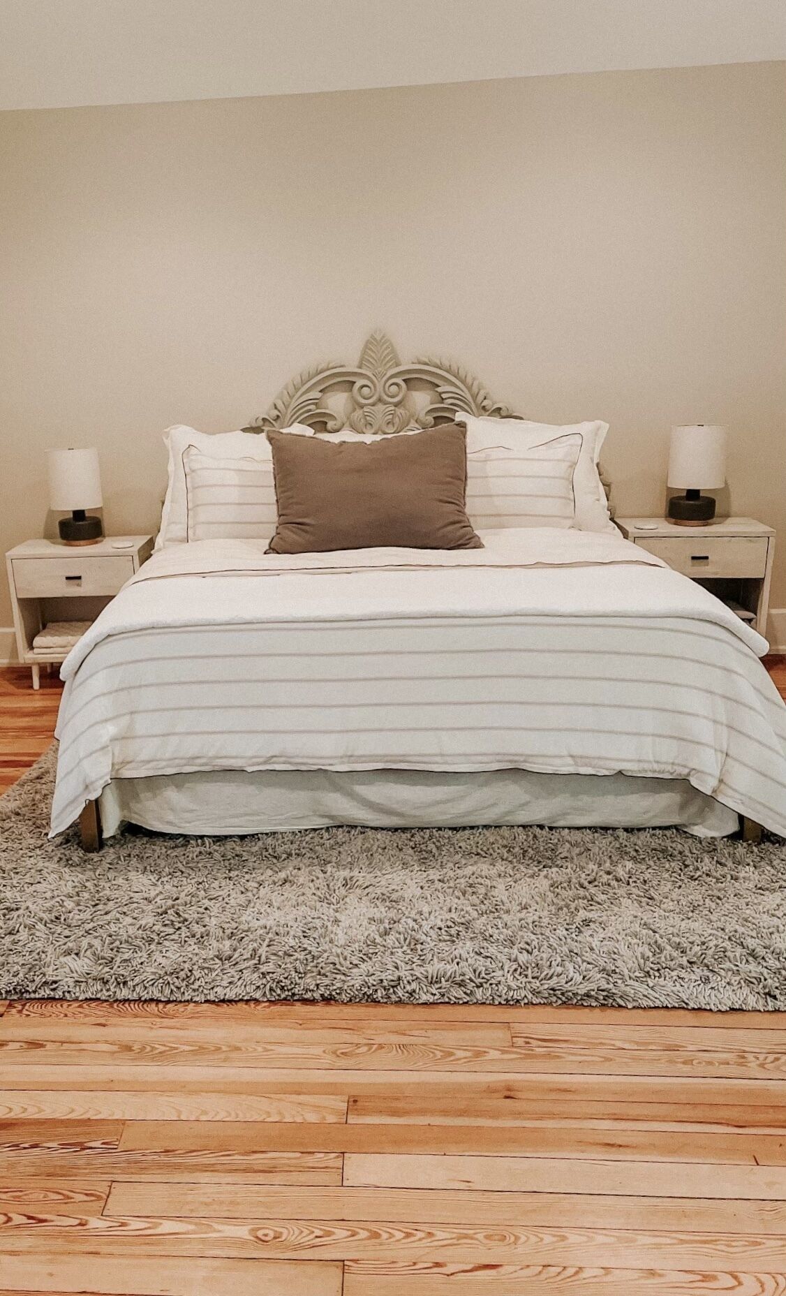A bed with a white and gray striped bedspread, a brown pillow, and white pillows. There are two bedside tables with lamps. The room has a wooden floor and a gray shag rug.