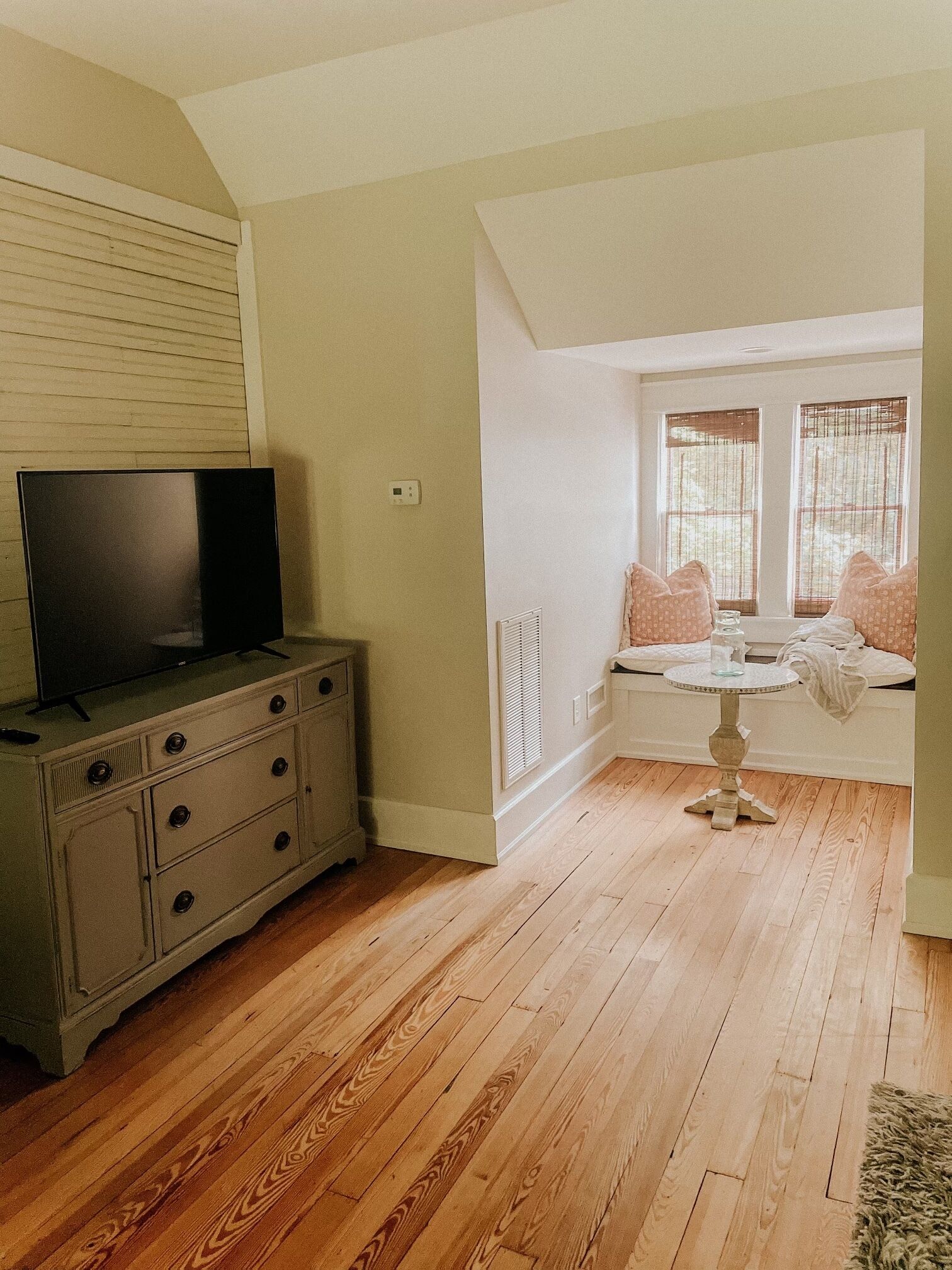 A room with a wooden floor, a dresser, a television on top, a window with blinds, and a small round table with a glass.
