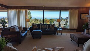 A living room with a view of the ocean, furnished with a sofa, two armchairs, and a coffee table. There are two lamps on side tables, and a tripod with a camera on the right side. - Panoramic Water Views, Retro Vibe, EV Charger, (Port Angeles)