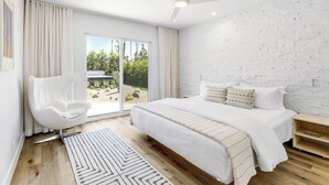 Een moderne slaapkamer met een groot bed, een witte stoel, een plafondventilator en uitzicht op groen.