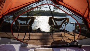 Ein Zelt mit zwei Stühlen, die auf ein Gewässer ausgerichtet sind, einem Tisch und einem Bett mit Decken.