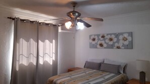 Une chambre à coucher avec un lit, un ventilateur de plafond, une fenêtre avec des rideaux et un tableau représentant des fleurs accroché au mur.