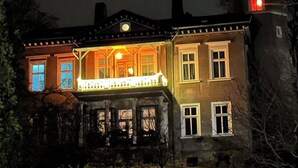 Ein zweistöckiges Haus mit Balkon und einem beleuchteten Fenster, umgeben von Bäumen.