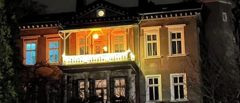 Una casa de dos pisos con un balcón y una ventana iluminada, rodeada de árboles.