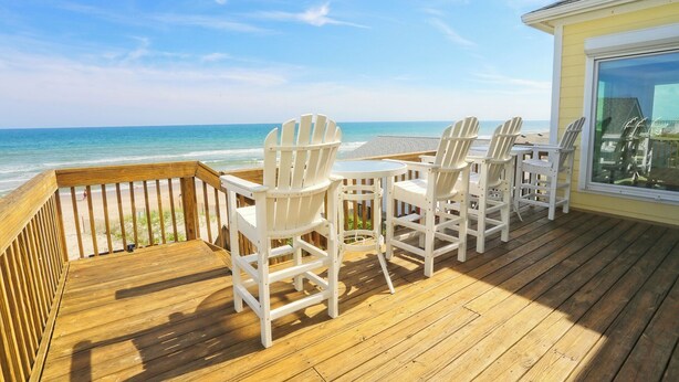 Een houten terras met witte Adirondack-stoelen en een tafel, met uitzicht op een strand en oceaan.