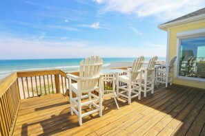 A wooden deck with white Adirondack chairs and a table, overlooking a beach and ocean.