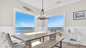 Een eetruimte met een houten tafel en stoelen, grote ramen met uitzicht op de oceaan, een kroonluchter en een wit metalen schap met keukenbenodigdheden.