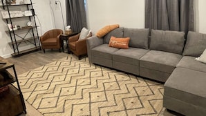 A modern living room with a grey sofa, two armchairs, a patterned rug, a bookshelf, and a window with curtains. - Cozy 2 Bed (Sterling)