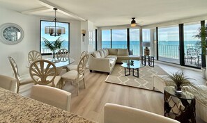 A spacious living room with a large sofa, a dining area with a glass table, and a view of the ocean through large windows. - Spectacular, spacious, renovated corner unit #501 with large wrap around balcony (Garden City)