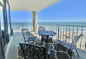 Un balcon avec une table et des chaises, donnant sur une plage où l’on peut voir des gens et des parasols.