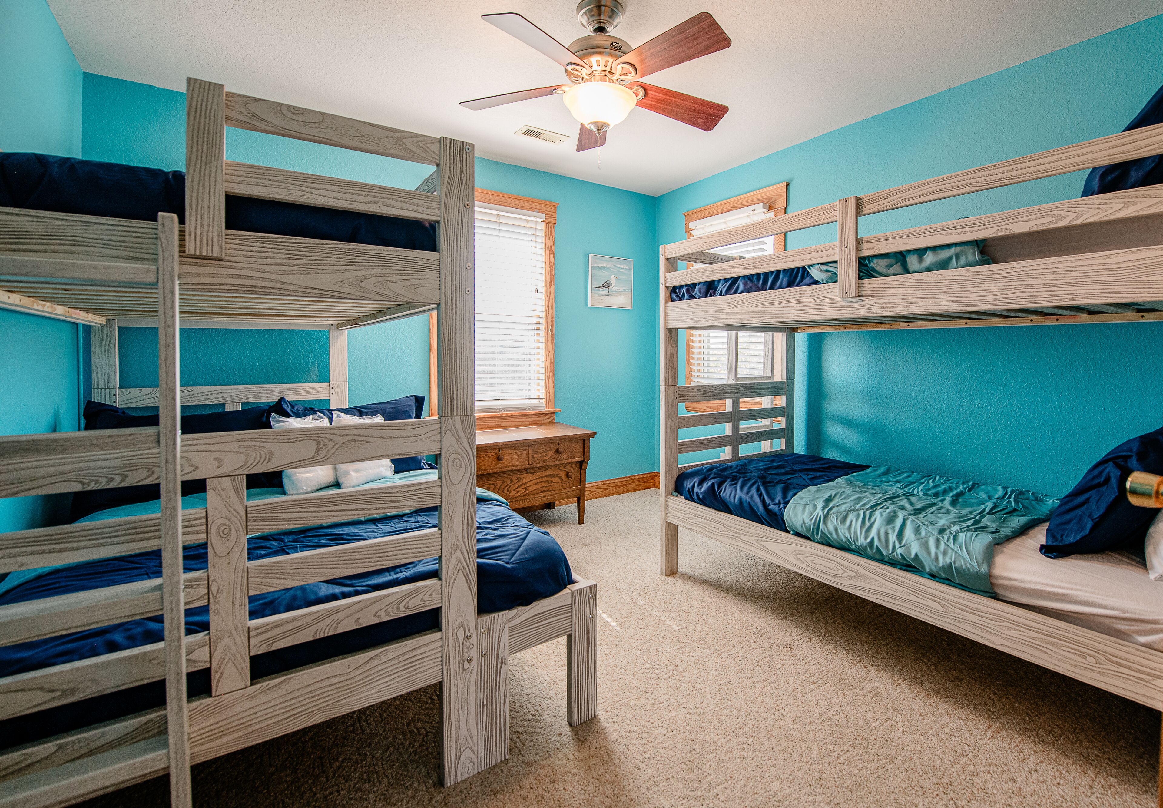 A room with bunk beds, a ceiling fan, a dresser, and a window with blinds.