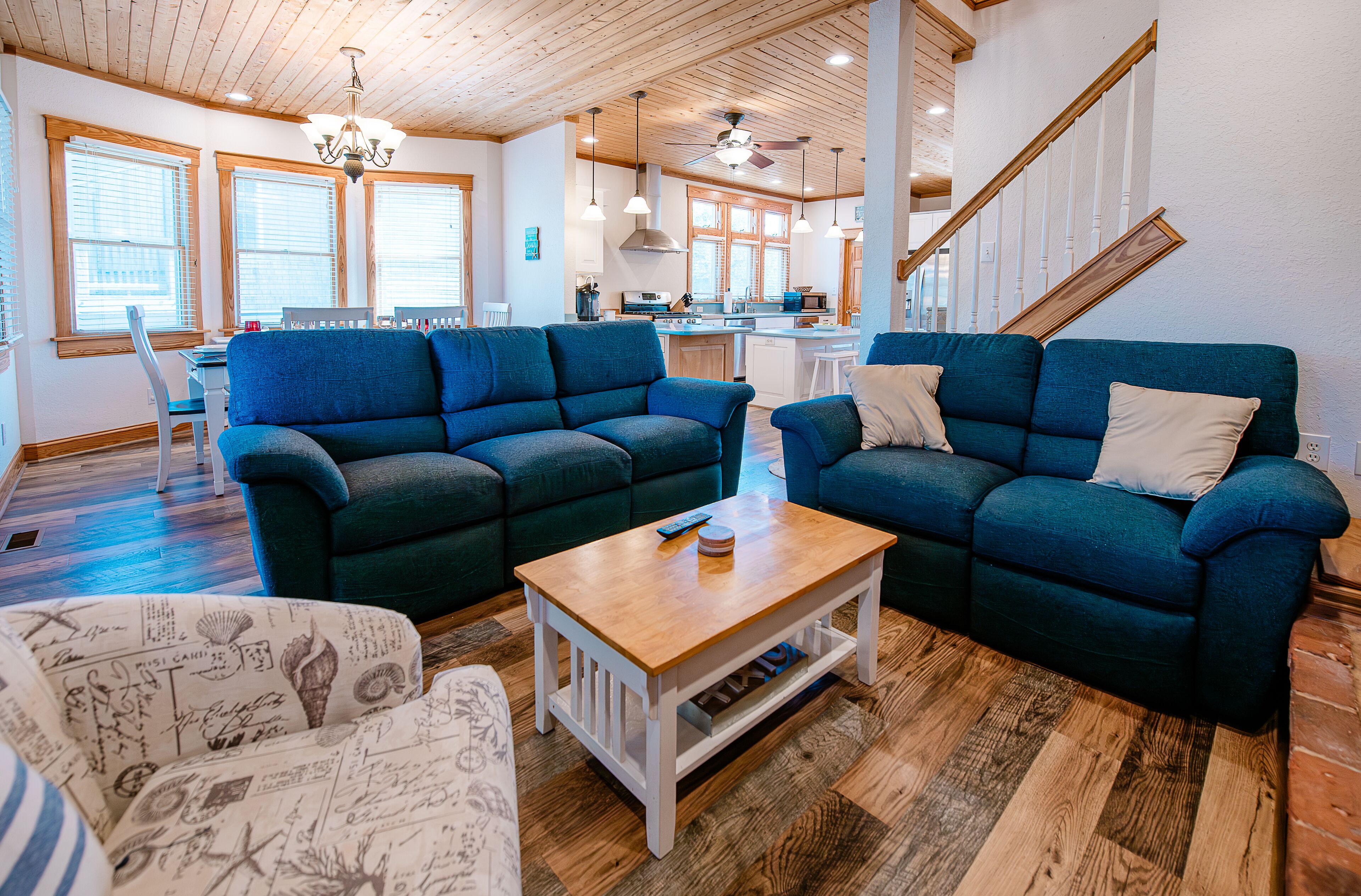 Un vaste salon comprenant un canapé d’angle bleu, une table basse en bois et un fauteuil à motifs. On aperçoit une cuisine à l’arrière-plan, avec des meubles de rangement en bois et un coin repas.