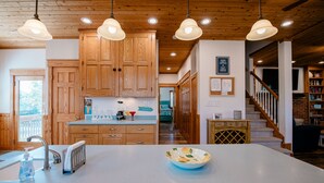 Une cuisine avec des meubles en bois, un grand îlot central doté d’un évier, des luminaires suspendus et une vue sur un escalier menant à l’espace de vie.