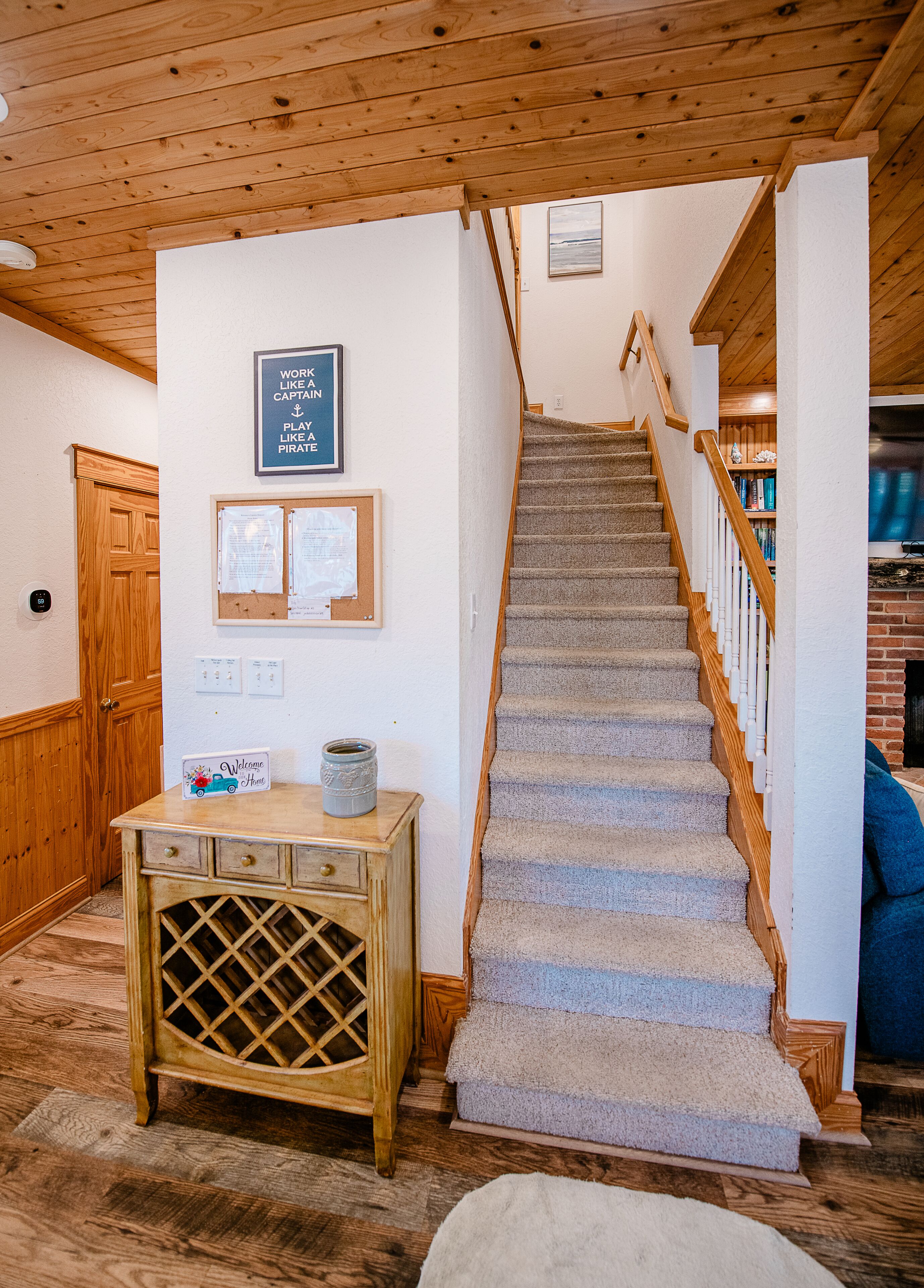 A staircase with wooden railings leading to a second floor, a wooden cabinet with a welcome sign, and a bulletin board with notices.